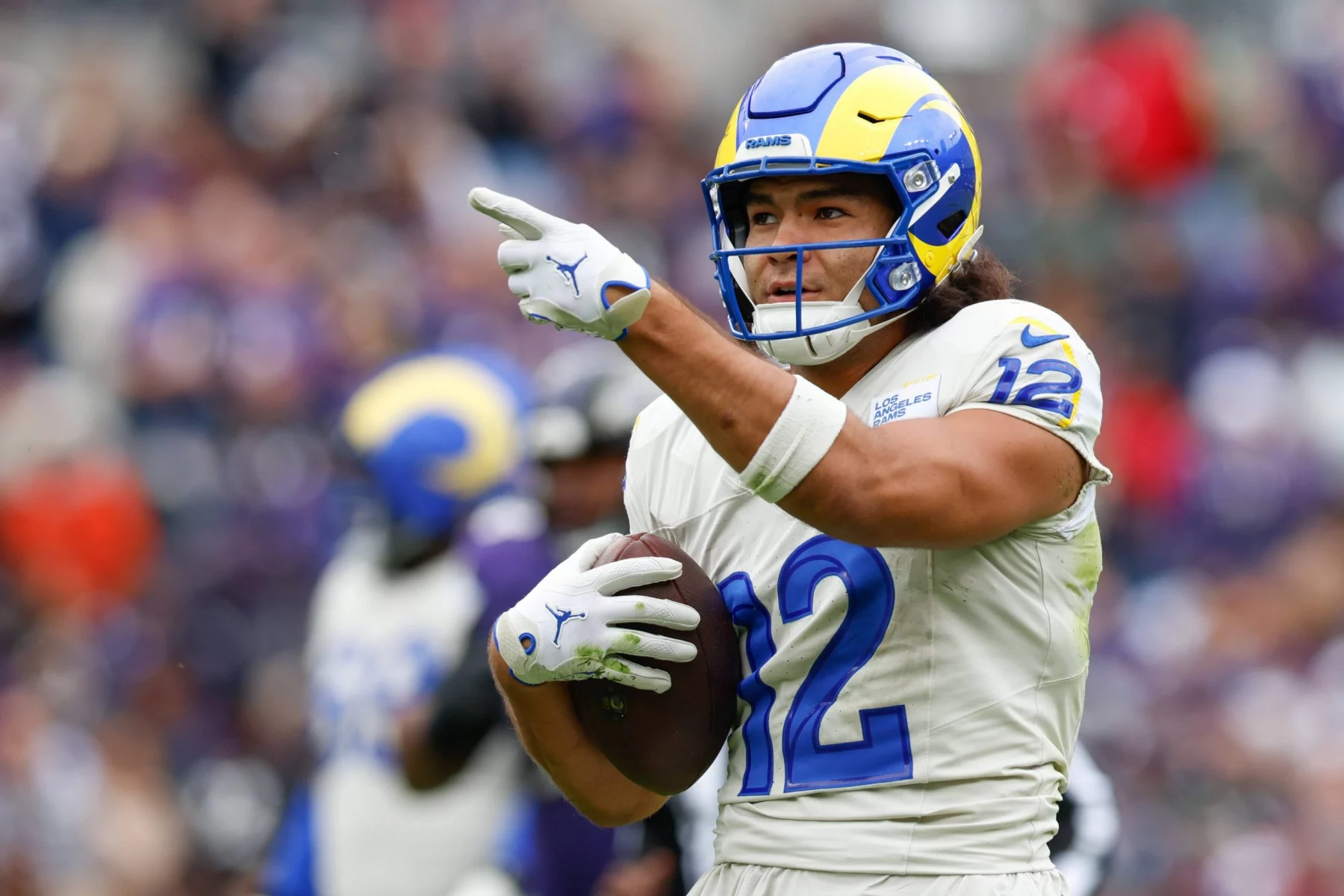 Fantasy Football - NFL Playoff Picture - Puka Nacua, Wide Receiver der Rams, in einer weiß-blauen Uniform und mit Helm, hält einen Football im rechten Arm und zeigt mit der linken Hand. Der Hintergrund ist unscharf und zeigt ein Publikum in einem Stadion. Diese Beschreibung wurde mit der FootballR KI automatisch generiert.