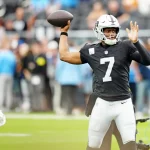 Geno Smith, der in einem schwarzen Trikot mit der Nummer 7 und einem silbernen Helm Quarterback Leistungen zeigt, befindet sich während der Saison 2025 mitten im Wurf. Ein anderer Mannschaftskamerad steht in der Nähe vor einer verschwommenen Menge und einem grünen Feld. Diese Beschreibung wurde mit der FootballR KI automatisch generiert.