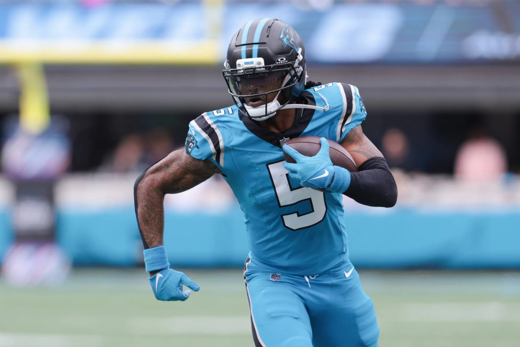 Rico Dowdle in einem blauen Trikot der Carolina Panthers mit der Nummer 5 läuft während eines Spiels mit dem Ball. Chuba Hubbard, einer der dynamischen RBs des Teams, trägt einen Helm und Handschuhe und blickt entschlossen nach vorne. Das Stadion und das Spielfeld verschwimmen im Hintergrund. Diese Beschreibung wurde mit der FootballR KI automatisch generiert.