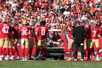 Eine Gruppe von Footballspielern der San Francisco 49ers, darunter Fred Warner in roten Trikots, versammelt sich um einen verletzten Mannschaftskameraden, der während der Saison auf einem Krankenwagen vom Spielfeld gebracht wird, während Trainer, Mitarbeiter und Fans von der Tribüne aus zusehen. Diese Beschreibung wurde mit der FootballR KI automatisch generiert.