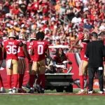 Eine Gruppe von Footballspielern der San Francisco 49ers, darunter Fred Warner in roten Trikots, versammelt sich um einen verletzten Mannschaftskameraden, der während der Saison auf einem Krankenwagen vom Spielfeld gebracht wird, während Trainer, Mitarbeiter und Fans von der Tribüne aus zusehen. Diese Beschreibung wurde mit der FootballR KI automatisch generiert.
