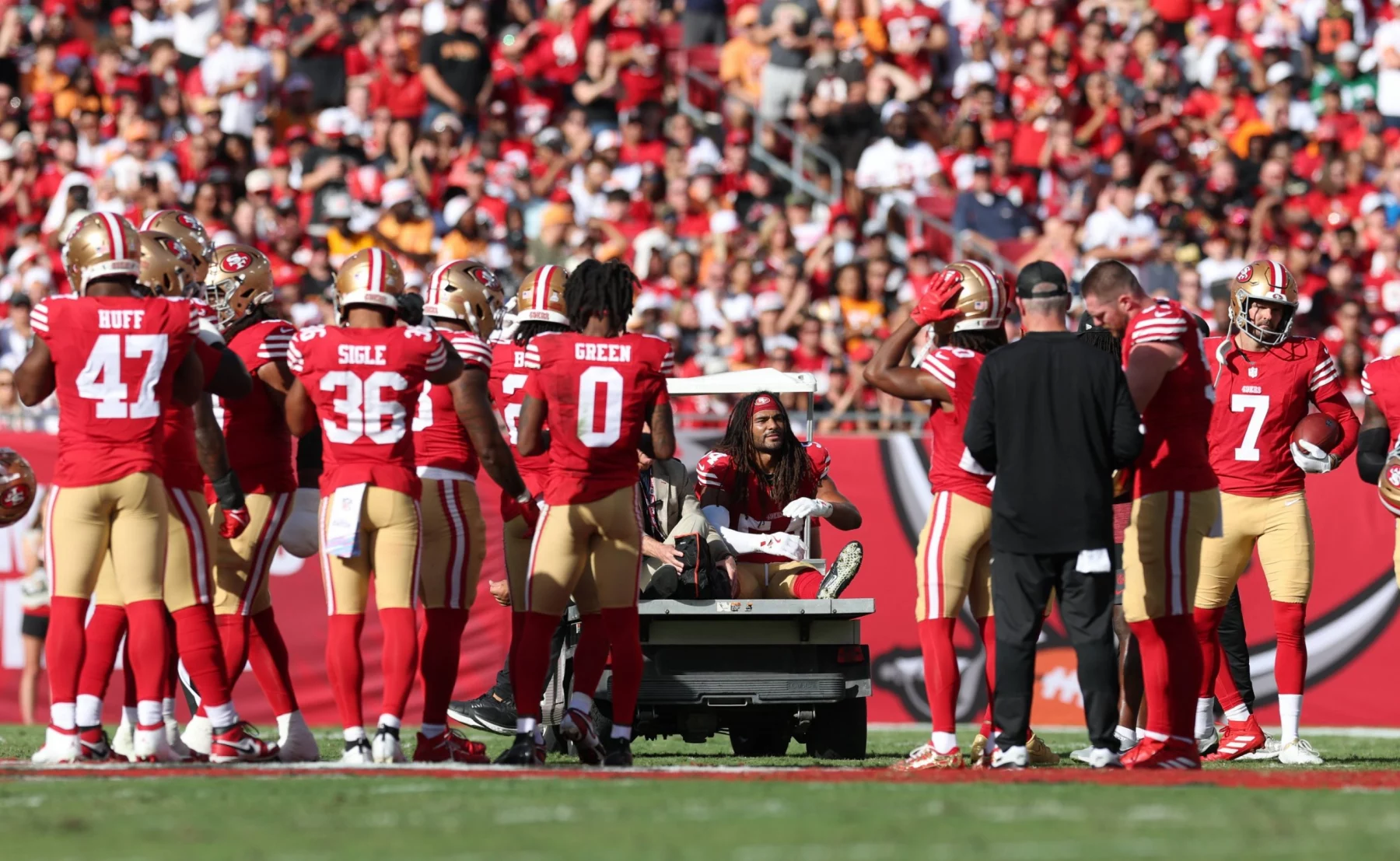 Eine Gruppe von Footballspielern der San Francisco 49ers, darunter Fred Warner in roten Trikots, versammelt sich um einen verletzten Mannschaftskameraden, der während der Saison auf einem Krankenwagen vom Spielfeld gebracht wird, während Trainer, Mitarbeiter und Fans von der Tribüne aus zusehen. Diese Beschreibung wurde mit der FootballR KI automatisch generiert.