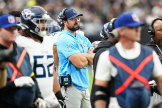 Titans-Cheftrainer Brian Callahan in hellblauem Hemd und Mütze steht während eines Spiels mit verschränkten Armen an der Seitenlinie, umgeben von Spielern in weißen und blauen Uniformen und fleißigem Personal, mit einer lebhaften Stadionmenge im Hintergrund. Diese Beschreibung wurde mit der FootballR KI automatisch generiert.