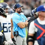 Titans-Cheftrainer Brian Callahan in hellblauem Hemd und Mütze steht während eines Spiels mit verschränkten Armen an der Seitenlinie, umgeben von Spielern in weißen und blauen Uniformen und fleißigem Personal, mit einer lebhaften Stadionmenge im Hintergrund. Diese Beschreibung wurde mit der FootballR KI automatisch generiert.
