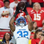 Detroit Lions-Safety Brian Branch, der ein weißes Trikot mit blauen Akzenten und der Nummer 32 trägt, fängt einen Football mit beiden Händen. NFL-Fans in roten und weißen Hemden, möglicherweise Chiefs-Anhänger, schauen aus dem unscharfen Hintergrund zu. Diese Beschreibung wurde mit der FootballR KI automatisch generiert.