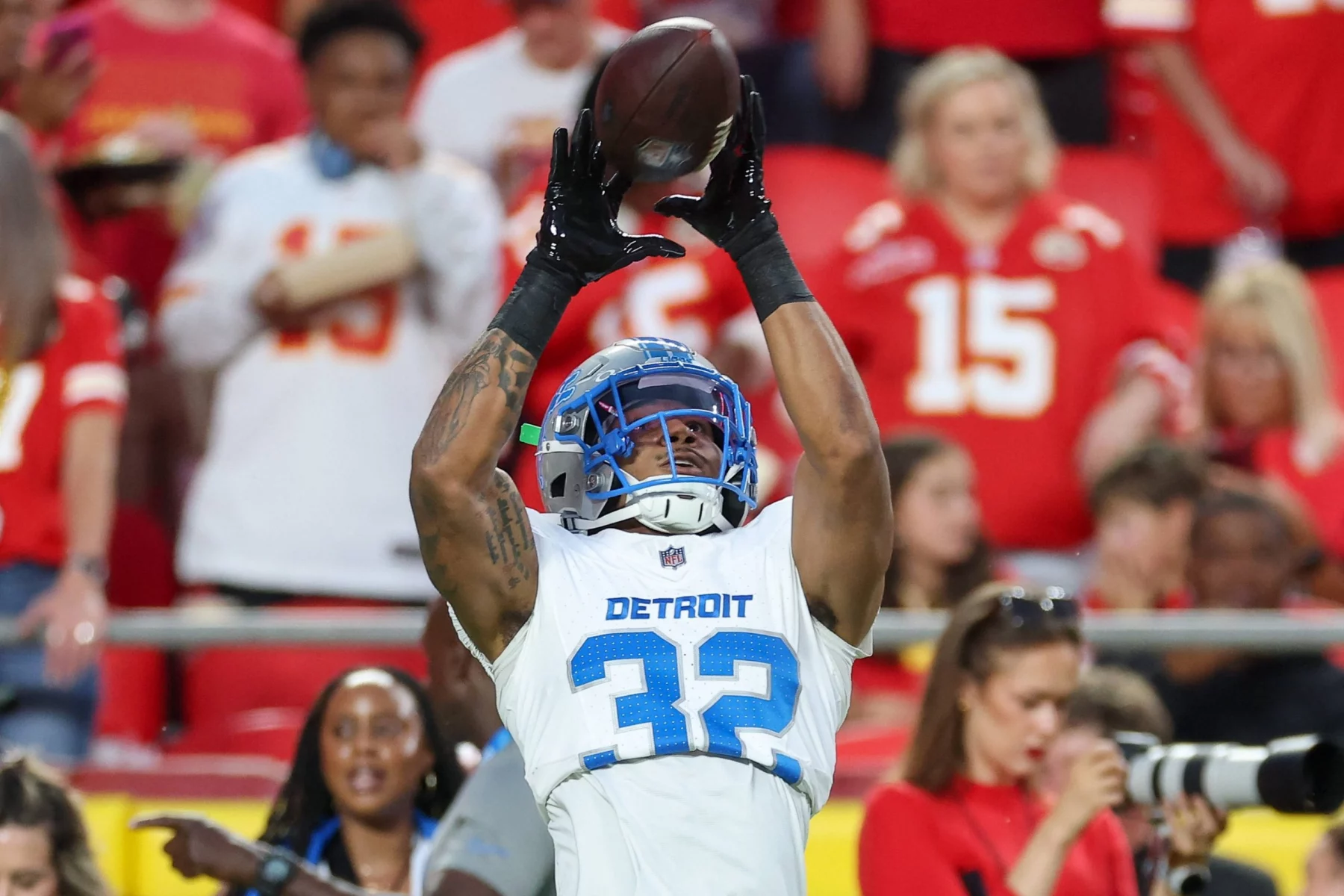 Detroit Lions-Safety Brian Branch, der ein weißes Trikot mit blauen Akzenten und der Nummer 32 trägt, fängt einen Football mit beiden Händen. NFL-Fans in roten und weißen Hemden, möglicherweise Chiefs-Anhänger, schauen aus dem unscharfen Hintergrund zu. Diese Beschreibung wurde mit der FootballR KI automatisch generiert.