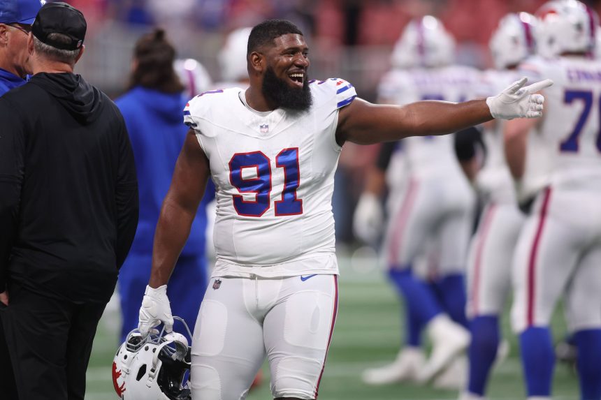 Ein lächelnder Bills-Footballspieler mit der Nummer 91 - Ed Oliver - steht in einer weißen Uniform mit blauen und roten Akzenten auf dem Feld, hält seinen Helm und gestikuliert. Andere Spieler und Trainer in Bills-Uniformen sind im unscharfen Hintergrund zu sehen. Diese Beschreibung wurde mit der FootballR KI automatisch generiert.