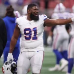 Ein lächelnder Bills-Footballspieler mit der Nummer 91 - Ed Oliver - steht in einer weißen Uniform mit blauen und roten Akzenten auf dem Feld, hält seinen Helm und gestikuliert. Andere Spieler und Trainer in Bills-Uniformen sind im unscharfen Hintergrund zu sehen. Diese Beschreibung wurde mit der FootballR KI automatisch generiert.