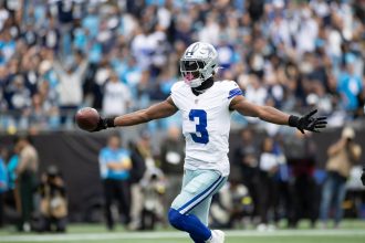 George Pickens, Dallas Cowboys, der die Trikotnummer 3 trägt, feiert mit ausgestreckten Armen, während er einen Football in der Hand hält, umgeben von einer unscharfen Menschenmenge in einem Stadion - vielleicht nach einem großen Spielzug oder einer gerüchteweisen Vertragsverlängerung mit den Cowboys. Diese Beschreibung wurde mit der FootballR KI automatisch generiert.