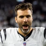 Joe Flacco - Ein Footballspieler in einer weiß-schwarzen Uniform steht auf dem Spielfeld, den Mund geöffnet, als würde er schreien. Er hat kurzes braunes Haar, einen Bart und sieht verschwitzt aus, ähnlich wie Joe Flacco. Der unscharfe Hintergrund zeigt ein volles Stadion. Diese Beschreibung wurde mit der FootballR KI automatisch generiert.