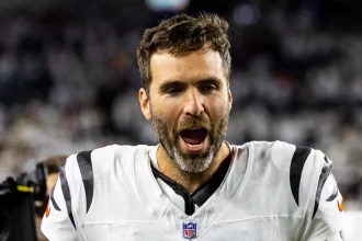 Joe Flacco - Ein Footballspieler in einer weiß-schwarzen Uniform steht auf dem Spielfeld, den Mund geöffnet, als würde er schreien. Er hat kurzes braunes Haar, einen Bart und sieht verschwitzt aus, ähnlich wie Joe Flacco. Der unscharfe Hintergrund zeigt ein volles Stadion. Diese Beschreibung wurde mit der FootballR KI automatisch generiert.