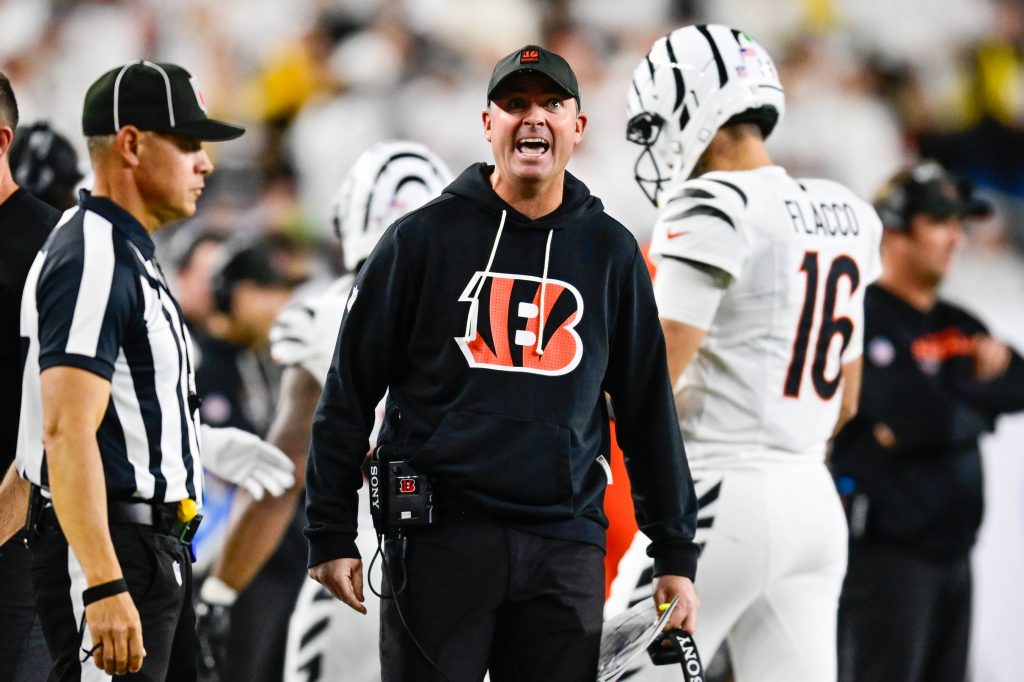 Ein Footballtrainer in einem schwarzen Bengals-Kapuzenpulli, wahrscheinlich Zac Taylor, schreit an der Seitenlinie und hält ein Spielblatt in der Hand. In der Nähe steht ein Schiedsrichter in einem gestreiften Hemd und einer Kappe. Im Hintergrund läuft ein Spieler in einem weißen Bengals-Trikot mit der Nummer 16 davon. Diese Beschreibung wurde mit der FootballR KI automatisch generiert.