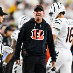 Ein Footballtrainer in einem schwarzen Bengals-Kapuzenpulli, wahrscheinlich Zac Taylor, schreit an der Seitenlinie und hält ein Spielblatt in der Hand. In der Nähe steht ein Schiedsrichter in einem gestreiften Hemd und einer Kappe. Im Hintergrund läuft ein Spieler in einem weißen Bengals-Trikot mit der Nummer 16 davon. Diese Beschreibung wurde mit der FootballR KI automatisch generiert.