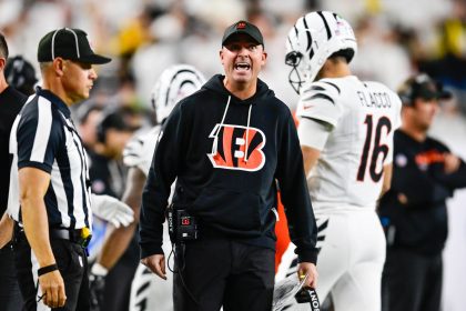 Ein Footballtrainer in einem schwarzen Bengals-Kapuzenpulli, wahrscheinlich Zac Taylor, schreit an der Seitenlinie und hält ein Spielblatt in der Hand. In der Nähe steht ein Schiedsrichter in einem gestreiften Hemd und einer Kappe. Im Hintergrund läuft ein Spieler in einem weißen Bengals-Trikot mit der Nummer 16 davon. Diese Beschreibung wurde mit der FootballR KI automatisch generiert.