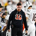 Ein Footballtrainer in einem schwarzen Bengals-Kapuzenpulli, wahrscheinlich Zac Taylor, schreit an der Seitenlinie und hält ein Spielblatt in der Hand. In der Nähe steht ein Schiedsrichter in einem gestreiften Hemd und einer Kappe. Im Hintergrund läuft ein Spieler in einem weißen Bengals-Trikot mit der Nummer 16 davon. Diese Beschreibung wurde mit der FootballR KI automatisch generiert.