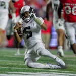 Woche 8 - Ein Football-Spieler in einem weißen Trikot mit der Nummer 5 - Dante Moore - rutscht auf einem Knie, hält einen Football und stützt sich mit der linken Hand ab. Spieler in roten Trikots verfolgen ihn auf dem grünen Feld - eine actiongeladene College-Football-Zusammenfassung von Woche 8. Diese Beschreibung wurde mit der FootballR KI automatisch generiert.