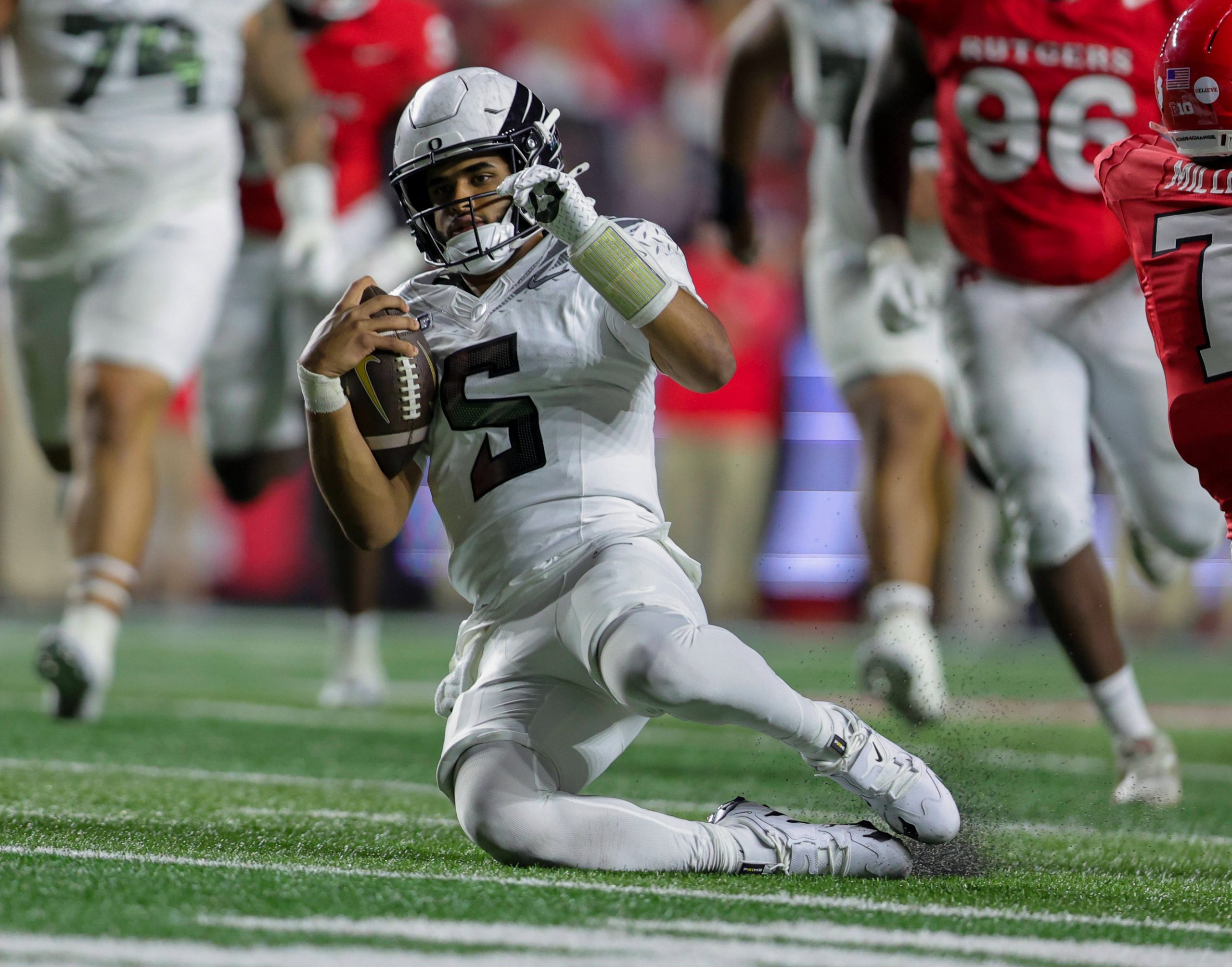 Woche 8 - Ein Football-Spieler in einem weißen Trikot mit der Nummer 5 - Dante Moore - rutscht auf einem Knie, hält einen Football und stützt sich mit der linken Hand ab. Spieler in roten Trikots verfolgen ihn auf dem grünen Feld - eine actiongeladene College-Football-Zusammenfassung von Woche 8. Diese Beschreibung wurde mit der FootballR KI automatisch generiert.