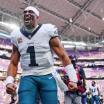 Woche 10 - Ein Footballspieler im weißen Trikot der Philadelphia Eagles mit der Nummer 1 ballt die Fäuste und jubelt auf dem Spielfeld während der NFL Woche 7. Das Stadion ist voll mit lila Sitzen und Menschen, während Kameras und Medien diesen elektrisierenden Moment festhalten. Diese Beschreibung wurde mit der FootballR KI automatisch generiert.