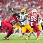 Micah Parsons, Green Bay Packers, in einem weiß-gelben Trikot versucht, an einem Spieler der Arizona Cardinals in Rot (#68) vorbeizulaufen, während ein anderer Spieler der Cardinals (#85) im Hintergrund während eines NFL-Spiels in Arizona auf einem Rasenfeld blockiert. Die Zuschauer sind unscharf. Diese Beschreibung wurde mit der FootballR KI automatisch generiert.