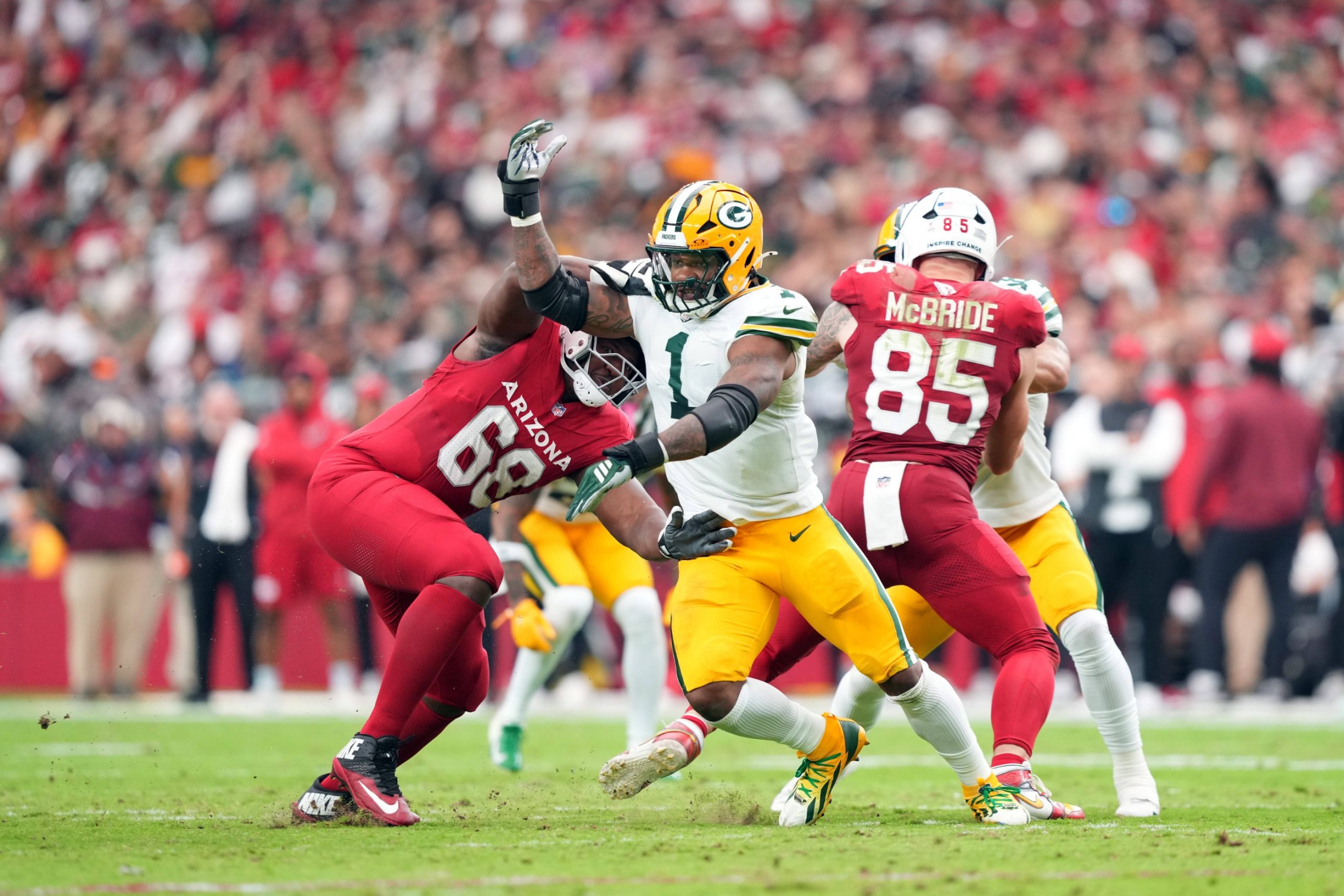Micah Parsons, Green Bay Packers, in einem weiß-gelben Trikot versucht, an einem Spieler der Arizona Cardinals in Rot (#68) vorbeizulaufen, während ein anderer Spieler der Cardinals (#85) im Hintergrund während eines NFL-Spiels in Arizona auf einem Rasenfeld blockiert. Die Zuschauer sind unscharf. Diese Beschreibung wurde mit der FootballR KI automatisch generiert.