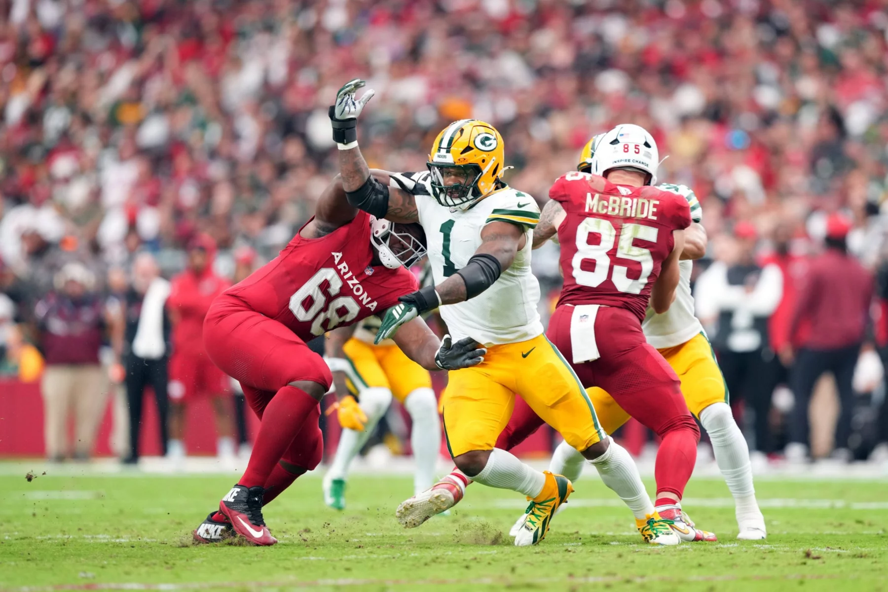 Micah Parsons, Green Bay Packers, in einem weiß-gelben Trikot versucht, an einem Spieler der Arizona Cardinals in Rot (#68) vorbeizulaufen, während ein anderer Spieler der Cardinals (#85) im Hintergrund während eines NFL-Spiels in Arizona auf einem Rasenfeld blockiert. Die Zuschauer sind unscharf. Diese Beschreibung wurde mit der FootballR KI automatisch generiert.