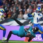 NFL Trade Deadline - Brian Thomas Jr. in einem blaugrünen Trikot fällt beim Fangen eines Footballs zu Boden, während zwei Spieler der Los Angeles Rams, von denen einer die Nummer 12 trägt, das Spiel aufmerksam verfolgen, während im unscharfen Hintergrund Trade-Angebote durch die Menge schwirren. Diese Beschreibung wurde mit der FootballR KI automatisch generiert.