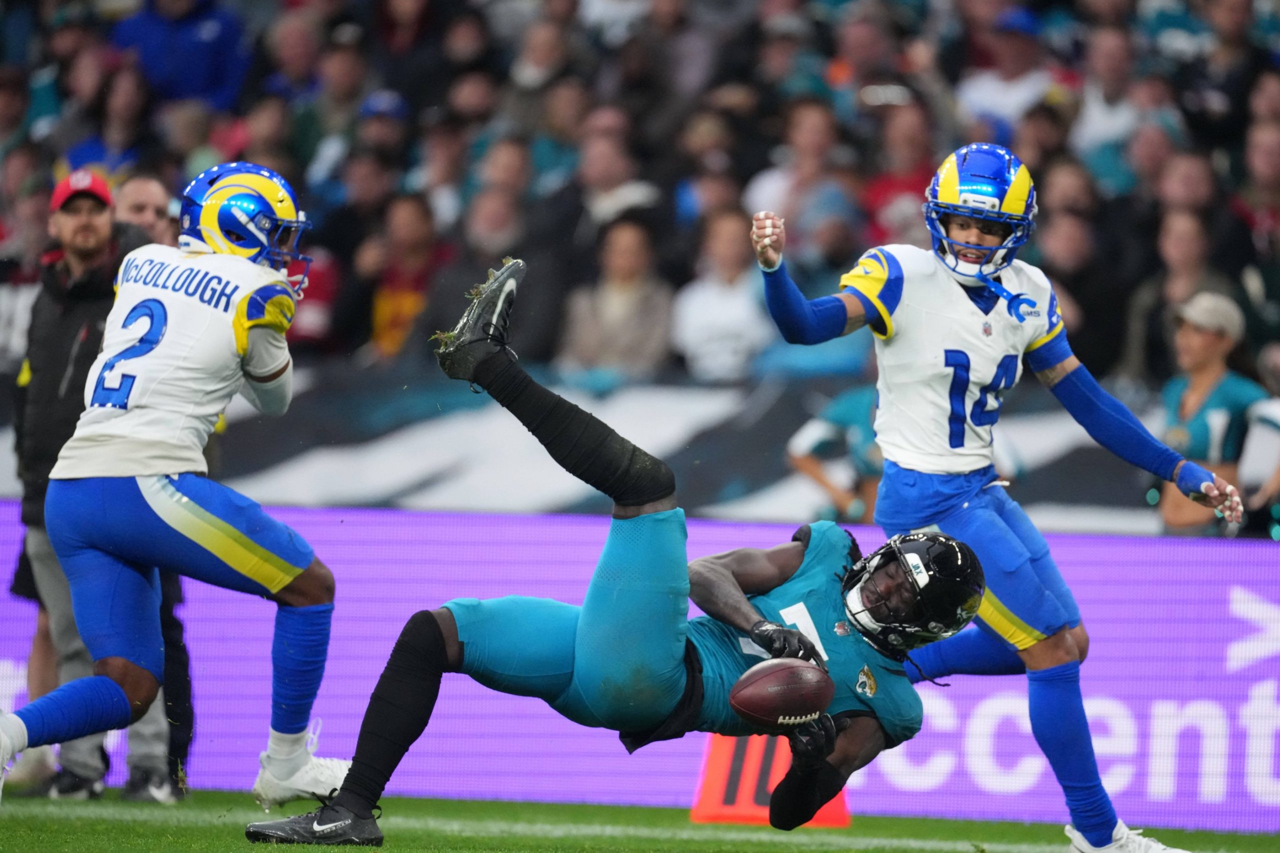 NFL Trade Deadline - Brian Thomas Jr. in einem blaugrünen Trikot fällt beim Fangen eines Footballs zu Boden, während zwei Spieler der Los Angeles Rams, von denen einer die Nummer 12 trägt, das Spiel aufmerksam verfolgen, während im unscharfen Hintergrund Trade-Angebote durch die Menge schwirren. Diese Beschreibung wurde mit der FootballR KI automatisch generiert.