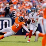 Ein Footballspieler in einer orangefarbenen Uniform stürzt sich auf Giants Quarterback Jaxson Dart, der den Ball hält und sich auf den Wurf vorbereitet. Ein weiterer orangefarbener Spieler ist unscharf im Vordergrund zu erkennen. Die Szene fängt einen spannungsgeladenen Moment ein, der an einen Kollaps der Giants erinnert. Diese Beschreibung wurde mit der FootballR KI automatisch generiert.