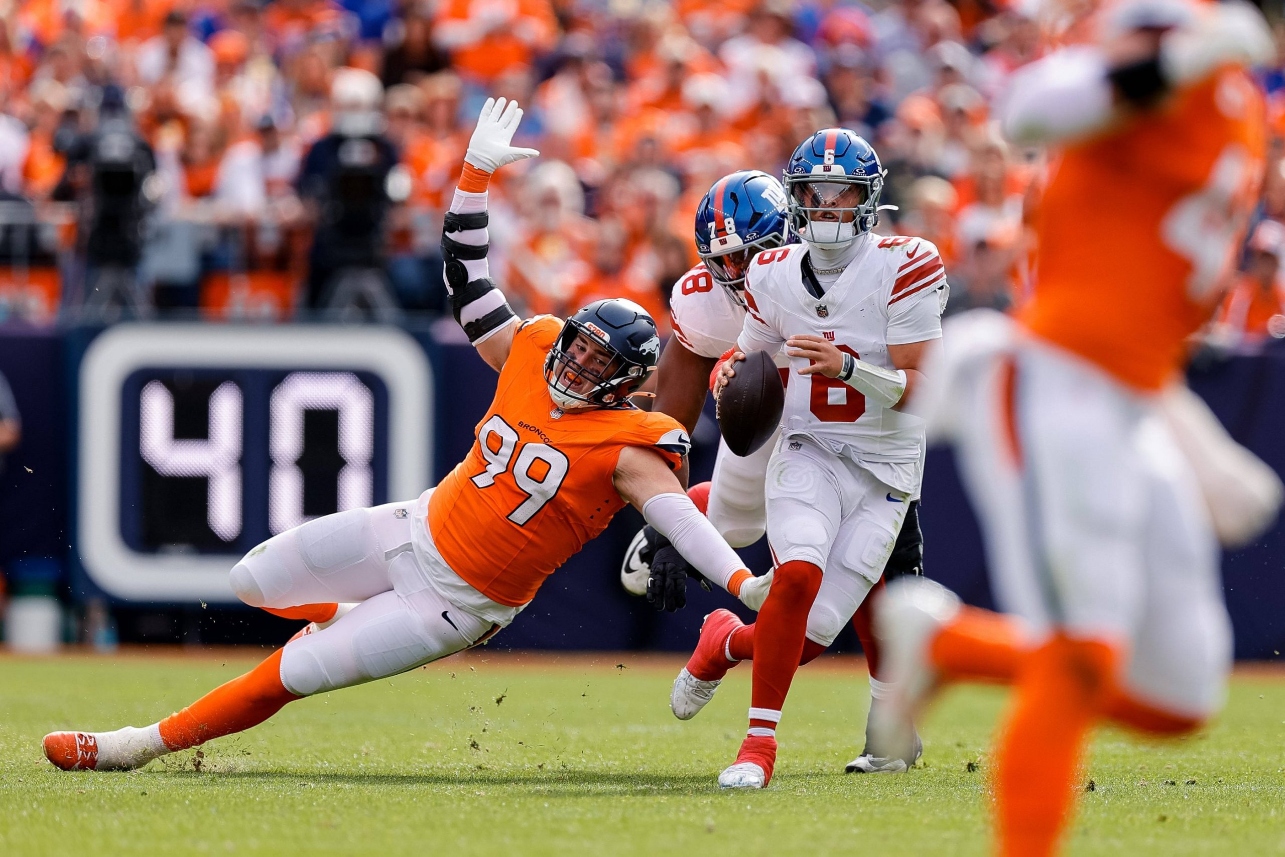 Ein Footballspieler in einer orangefarbenen Uniform stürzt sich auf Giants Quarterback Jaxson Dart, der den Ball hält und sich auf den Wurf vorbereitet. Ein weiterer orangefarbener Spieler ist unscharf im Vordergrund zu erkennen. Die Szene fängt einen spannungsgeladenen Moment ein, der an einen Kollaps der Giants erinnert. Diese Beschreibung wurde mit der FootballR KI automatisch generiert.