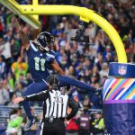 Ein Spieler der Seattle Seahawks, möglicherweise Smith-Njigba, hängt nach einem Tor mit einem Arm am gelben Torpfosten, die Beine weit gespreizt. Ein Schiedsrichter darunter trägt die Nummer "132". Jubelnde Fans füllen das Stadion; der Torpfosten ist in Regenbogenfarben und ein NFL-Logo gehüllt. Diese Beschreibung wurde mit der FootballR KI automatisch generiert.
