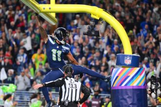 Ein Spieler der Seattle Seahawks, möglicherweise Smith-Njigba, hängt nach einem Tor mit einem Arm am gelben Torpfosten, die Beine weit gespreizt. Ein Schiedsrichter darunter trägt die Nummer "132". Jubelnde Fans füllen das Stadion; der Torpfosten ist in Regenbogenfarben und ein NFL-Logo gehüllt. Diese Beschreibung wurde mit der FootballR KI automatisch generiert.