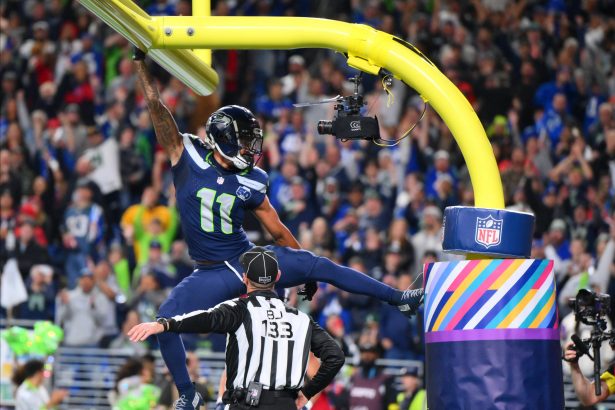 Woche 18 - Woche 11 - Ein Spieler der Seattle Seahawks, möglicherweise Smith-Njigba, hängt nach einem Tor mit einem Arm am gelben Torpfosten, die Beine weit gespreizt. Ein Schiedsrichter darunter trägt die Nummer "132". Jubelnde Fans füllen das Stadion; der Torpfosten ist in Regenbogenfarben und ein NFL-Logo gehüllt. Diese Beschreibung wurde mit der FootballR KI automatisch generiert.