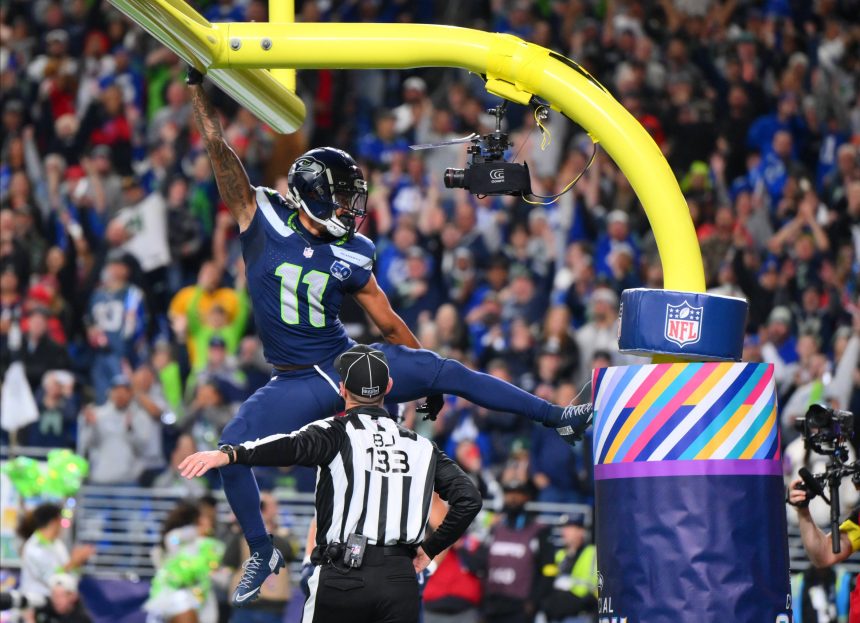 Ein Spieler der Seattle Seahawks, möglicherweise Smith-Njigba, hängt nach einem Tor mit einem Arm am gelben Torpfosten, die Beine weit gespreizt. Ein Schiedsrichter darunter trägt die Nummer "132". Jubelnde Fans füllen das Stadion; der Torpfosten ist in Regenbogenfarben und ein NFL-Logo gehüllt. Diese Beschreibung wurde mit der FootballR KI automatisch generiert.