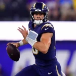 Justin Herbert in einem dunkelblauen Chargers-Trikot mit gelben Verzierungen und einem Blitz-Logo läuft mit dem Ball und schaut nach links. Er trägt einen Helm, hat ein Spielblatt am linken Handgelenk und wirkt inmitten der Stadionbeleuchtung und der Menge konzentriert. Diese Beschreibung wurde mit der FootballR KI automatisch generiert.