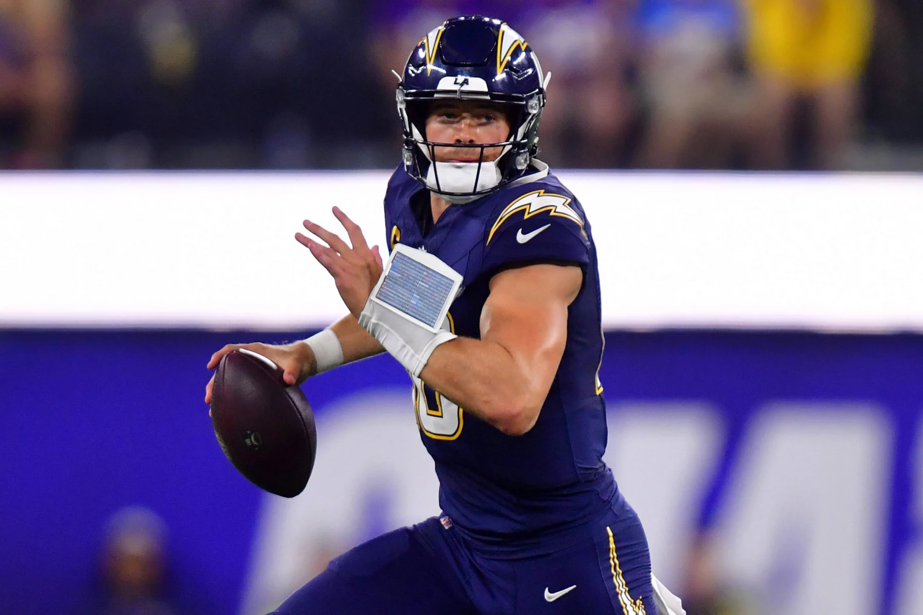 Justin Herbert in einem dunkelblauen Chargers-Trikot mit gelben Verzierungen und einem Blitz-Logo läuft mit dem Ball und schaut nach links. Er trägt einen Helm, hat ein Spielblatt am linken Handgelenk und wirkt inmitten der Stadionbeleuchtung und der Menge konzentriert. Diese Beschreibung wurde mit der FootballR KI automatisch generiert.