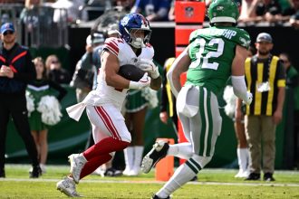 Cam Skattebo, New York Giants, in einer weißen Uniform läuft mit dem Ball, verfolgt von einem Spieler der Jets in Grün auf einem sonnigen Feld. Cheerleader und Offizielle sind zu sehen, während die Zuschauer von der Tribüne aus den Spielzug der Giants verfolgen. Diese Beschreibung wurde mit der FootballR KI automatisch generiert.