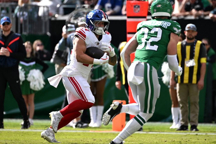 Cam Skattebo, New York Giants, in einer weißen Uniform läuft mit dem Ball, verfolgt von einem Spieler der Jets in Grün auf einem sonnigen Feld. Cheerleader und Offizielle sind zu sehen, während die Zuschauer von der Tribüne aus den Spielzug der Giants verfolgen. Diese Beschreibung wurde mit der FootballR KI automatisch generiert.