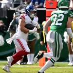 Cam Skattebo, New York Giants, in einer weißen Uniform läuft mit dem Ball, verfolgt von einem Spieler der Jets in Grün auf einem sonnigen Feld. Cheerleader und Offizielle sind zu sehen, während die Zuschauer von der Tribüne aus den Spielzug der Giants verfolgen. Diese Beschreibung wurde mit der FootballR KI automatisch generiert.