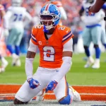 Broncos-Star-Cornerback Pat Surtain II in seinem orangefarbenen Trikot mit der Nummer 2 kniet auf dem Spielfeld, ballt die Fäuste und schreit vor der jubelnden Menge im Stadion, während Spieler in Blau und Weiß und ein Schiedsrichter in der Nähe stehen. Diese Beschreibung wurde mit der FootballR KI automatisch generiert.