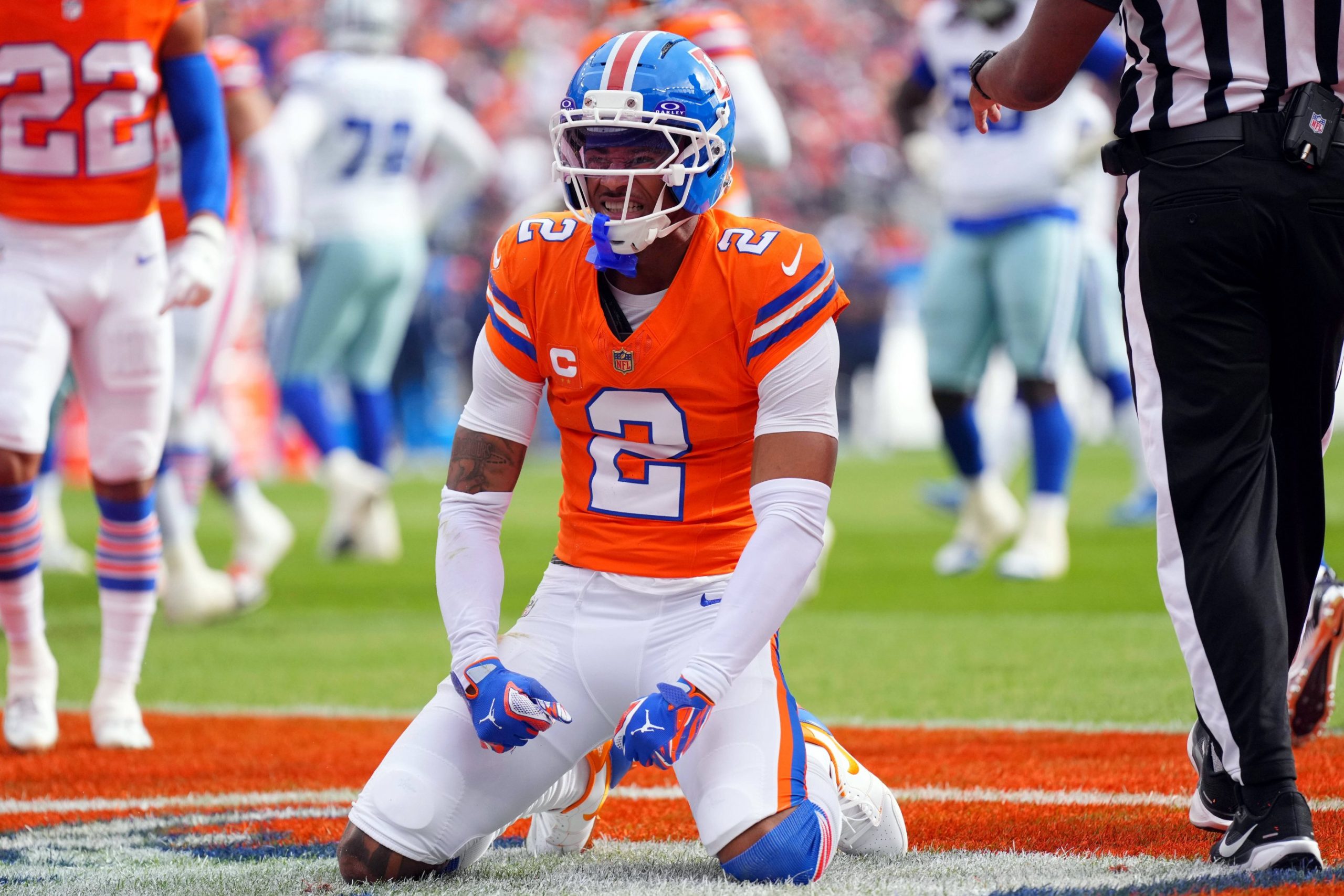 Broncos-Star-Cornerback Pat Surtain II in seinem orangefarbenen Trikot mit der Nummer 2 kniet auf dem Spielfeld, ballt die Fäuste und schreit vor der jubelnden Menge im Stadion, während Spieler in Blau und Weiß und ein Schiedsrichter in der Nähe stehen. Diese Beschreibung wurde mit der FootballR KI automatisch generiert.