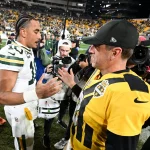 Zwei NFL-Spieler, Packers-Quarterback Jordan Love und Ex-Mentor Aaron Rodgers, unterhalten sich nach dem Spiel auf dem Spielfeld. Der eine trägt eine Green Bay-Uniform, der andere ein Steelers-Trikot. Die Fotografen halten den lebhaften Moment fest, während die Fans das hell erleuchtete Stadion füllen. Diese Beschreibung wurde mit der FootballR KI automatisch generiert.