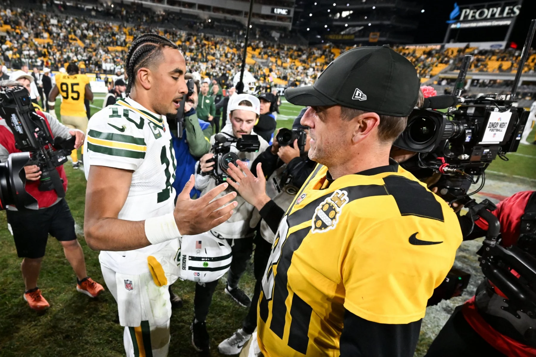 Zwei NFL-Spieler, Packers-Quarterback Jordan Love und Ex-Mentor Aaron Rodgers, unterhalten sich nach dem Spiel auf dem Spielfeld. Der eine trägt eine Green Bay-Uniform, der andere ein Steelers-Trikot. Die Fotografen halten den lebhaften Moment fest, während die Fans das hell erleuchtete Stadion füllen. Diese Beschreibung wurde mit der FootballR KI automatisch generiert.