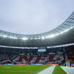 Das Berliner Olympiastadion ist voll mit Fans, die mit farbigen Karten ein Mosaik bilden, auf dem "NFL" in Rot und Weiß steht, umgeben von Schwarz und Gelb. Auf dem grünen Spielfeld befinden sich Endzonen, Torpfosten und eine Anzeigetafel unter einem wolkenverhangenen Himmel. Diese Beschreibung wurde mit der FootballR KI automatisch generiert.