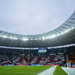 Das Berliner Olympiastadion ist voll mit Fans, die mit farbigen Karten ein Mosaik bilden, auf dem "NFL" in Rot und Weiß steht, umgeben von Schwarz und Gelb. Auf dem grünen Spielfeld befinden sich Endzonen, Torpfosten und eine Anzeigetafel unter einem wolkenverhangenen Himmel. Diese Beschreibung wurde mit der FootballR KI automatisch generiert.