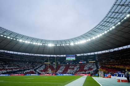Das Berliner Olympiastadion ist voll mit Fans, die mit farbigen Karten ein Mosaik bilden, auf dem "NFL" in Rot und Weiß steht, umgeben von Schwarz und Gelb. Auf dem grünen Spielfeld befinden sich Endzonen, Torpfosten und eine Anzeigetafel unter einem wolkenverhangenen Himmel. Diese Beschreibung wurde mit der FootballR KI automatisch generiert.