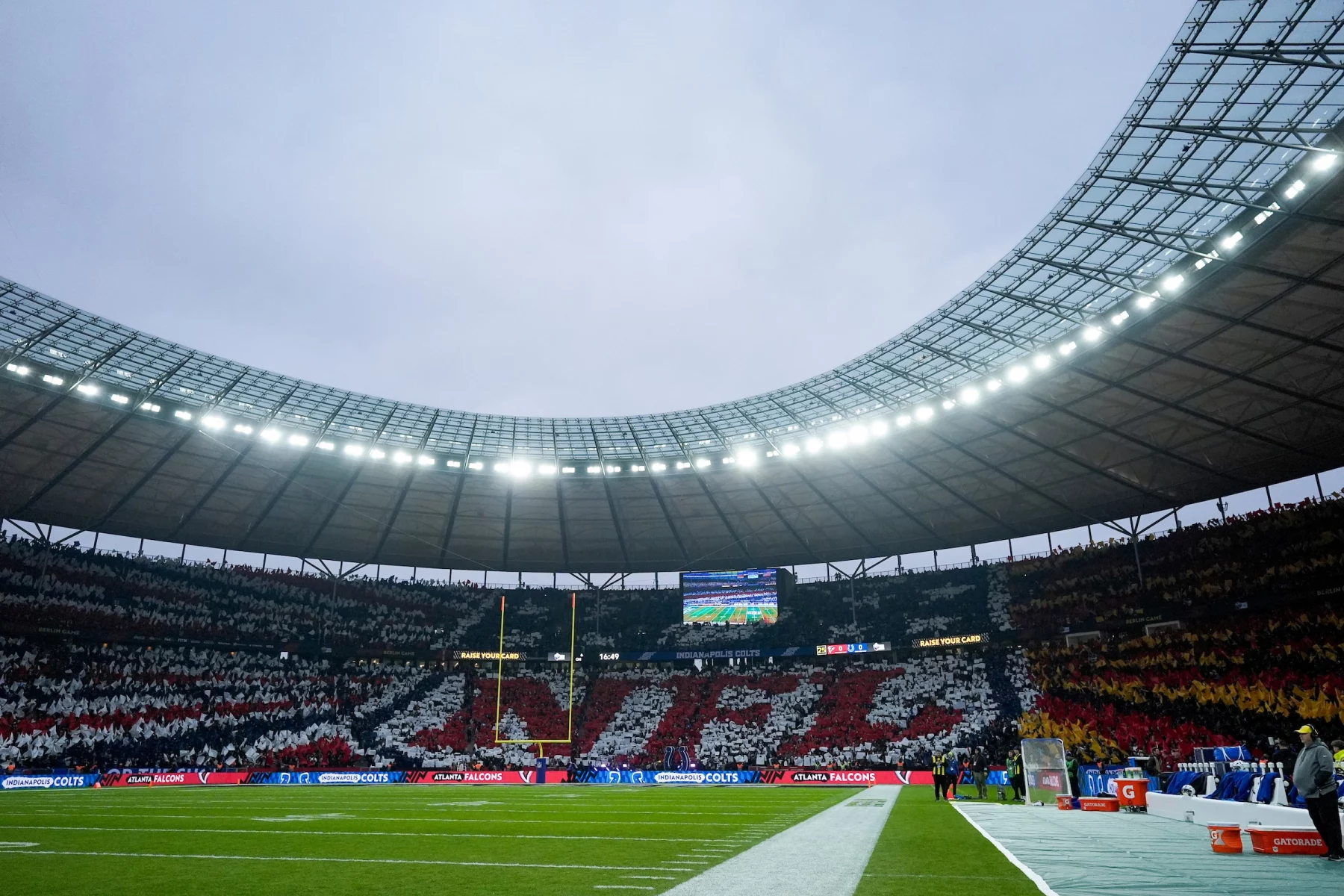 Das Berliner Olympiastadion ist voll mit Fans, die mit farbigen Karten ein Mosaik bilden, auf dem "NFL" in Rot und Weiß steht, umgeben von Schwarz und Gelb. Auf dem grünen Spielfeld befinden sich Endzonen, Torpfosten und eine Anzeigetafel unter einem wolkenverhangenen Himmel. Diese Beschreibung wurde mit der FootballR KI automatisch generiert.