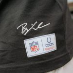 Eine Nahaufnahme einer Person, die ein schwarzes Hemd aus der Merchandise-Kollektion mit einer weißen Hose trägt. Das Hemd trägt Björn Werners Unterschrift über zwei Aufnähern: einem NFL-Logo-Aufnäher und einem Indianapolis Colts Mitchell & Ness-Aufnäher. Der Hintergrund ist unscharf. Diese Beschreibung wurde mit der FootballR KI automatisch generiert.