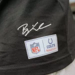Eine Nahaufnahme einer Person, die ein schwarzes Hemd aus der Merchandise-Kollektion mit einer weißen Hose trägt. Das Hemd trägt Björn Werners Unterschrift über zwei Aufnähern: einem NFL-Logo-Aufnäher und einem Indianapolis Colts Mitchell & Ness-Aufnäher. Der Hintergrund ist unscharf. Diese Beschreibung wurde mit der FootballR KI automatisch generiert.
