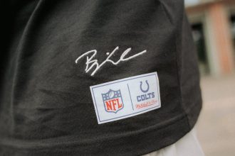 Eine Nahaufnahme einer Person, die ein schwarzes Hemd aus der Merchandise-Kollektion mit einer weißen Hose trägt. Das Hemd trägt Björn Werners Unterschrift über zwei Aufnähern: einem NFL-Logo-Aufnäher und einem Indianapolis Colts Mitchell & Ness-Aufnäher. Der Hintergrund ist unscharf. Diese Beschreibung wurde mit der FootballR KI automatisch generiert.