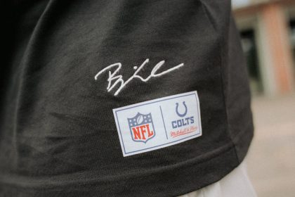 Eine Nahaufnahme einer Person, die ein schwarzes Hemd aus der Merchandise-Kollektion mit einer weißen Hose trägt. Das Hemd trägt Björn Werners Unterschrift über zwei Aufnähern: einem NFL-Logo-Aufnäher und einem Indianapolis Colts Mitchell & Ness-Aufnäher. Der Hintergrund ist unscharf. Diese Beschreibung wurde mit der FootballR KI automatisch generiert.